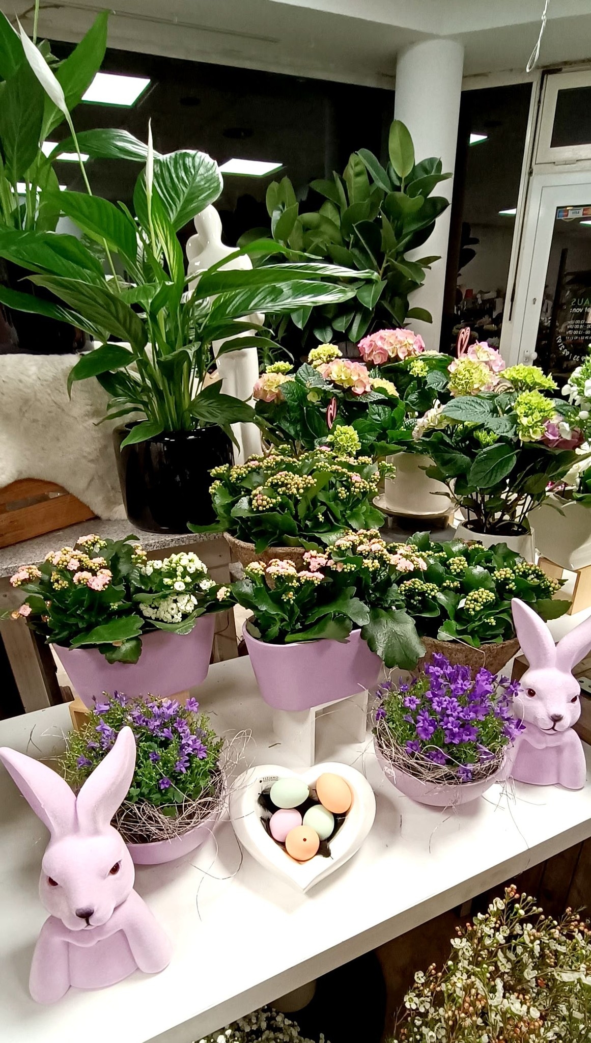 Frühlingshafte Osterdekoration in Pastelltönen mit lila Osterhasen-Figuren, violetten Glockenblumen, fliederfarbenen Pflanztöpfen und bunten Deko-Eiern in einer Herzschale.