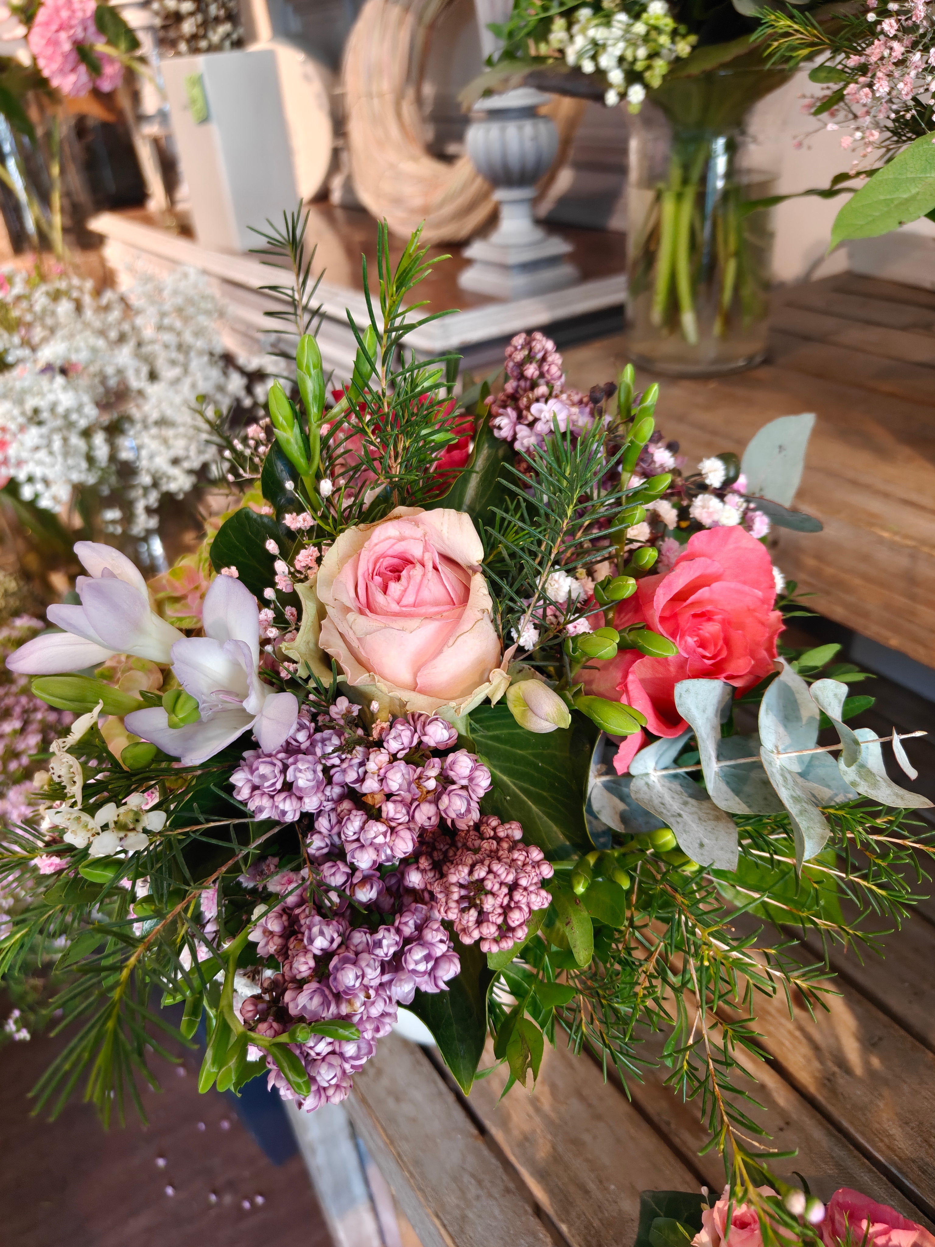 Bunter Blumenstrauß mit rosa Rosen, fliederfarbenem Flieder, weißen Freesien und Eukalyptusblättern auf einem Holztisch.